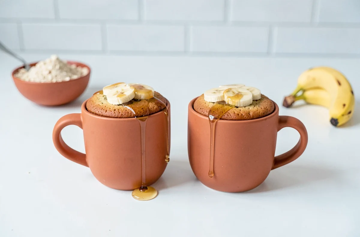 Deux mugcakes banane sans oeuf dans des mugs terracotta garnis de rondelles de banane et d'un filet de miel sur un comptoir blanc